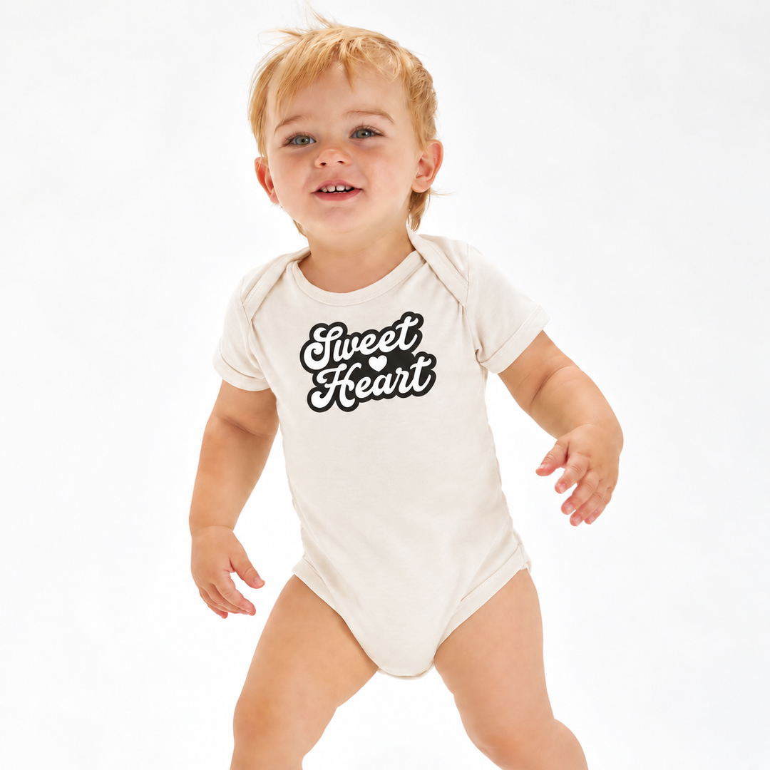 Baby draagt naturel rompertje met Sweet Heart opdruk.