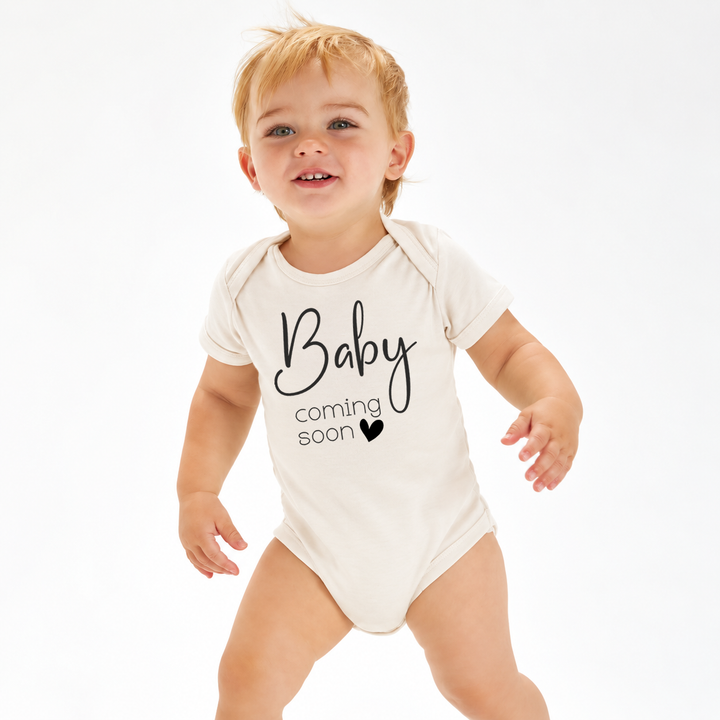 Baby draagt naturel romper met zwarte tekst Baby Coming Soon.