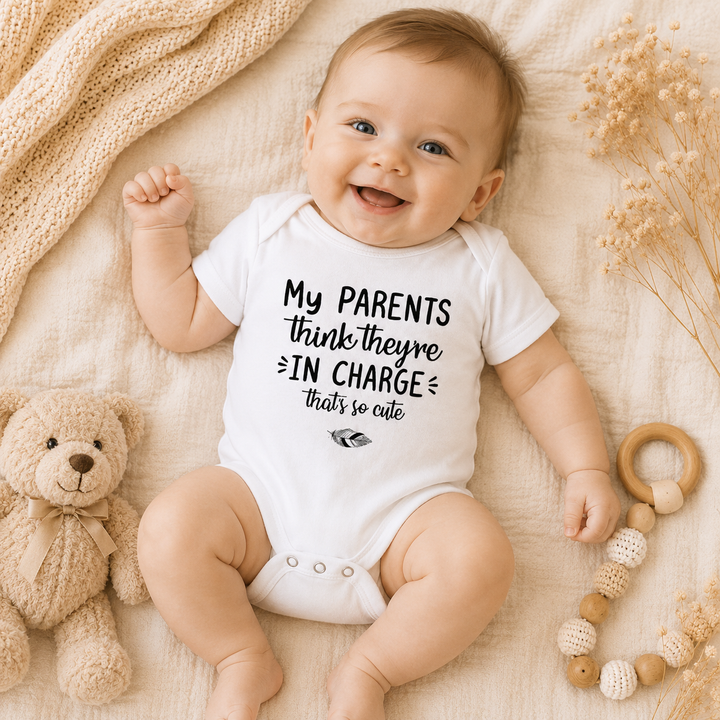 Lachende baby draagt witte rompertje met tekst “My parents think they’re in charge that’s so cute” – grappig rompertje met tekst als kraamcadeau en zwangerschapsaankondiging