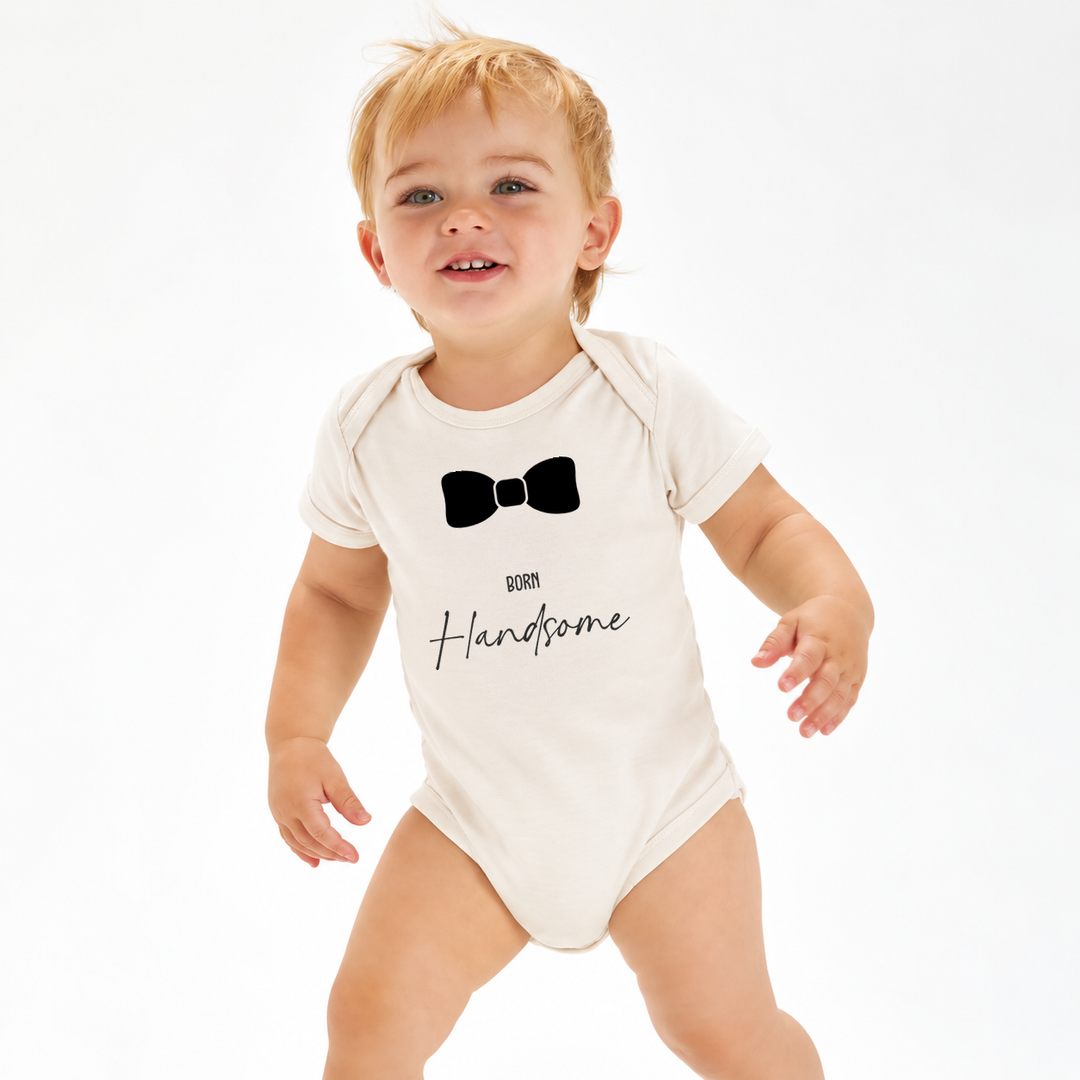 Baby draagt naturel romper jongen met Born Handsome tekst en zwarte strik.
