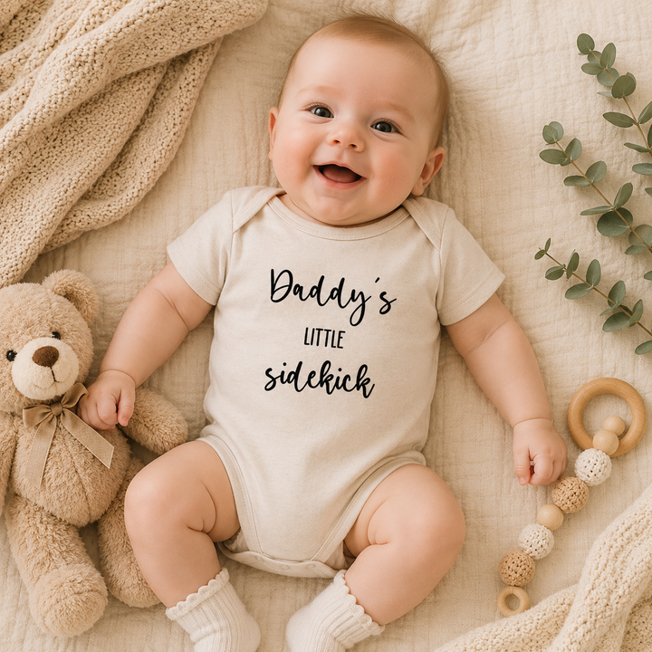 Blije baby in beige rompertje met tekst “Daddy’s little sidekick” op bed, origineel baby cadeau en zwangerschapsaankondiging rompertje.
