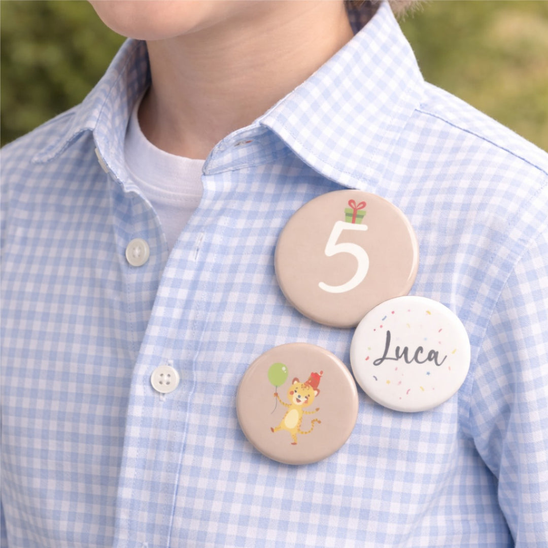 Drie gepersonaliseerde verjaardagsbuttons met het getal 5, de naam Luca en een tijgerillustratie, gedragen op een lichtblauw overhemd door een kind tijdens een verjaardag.