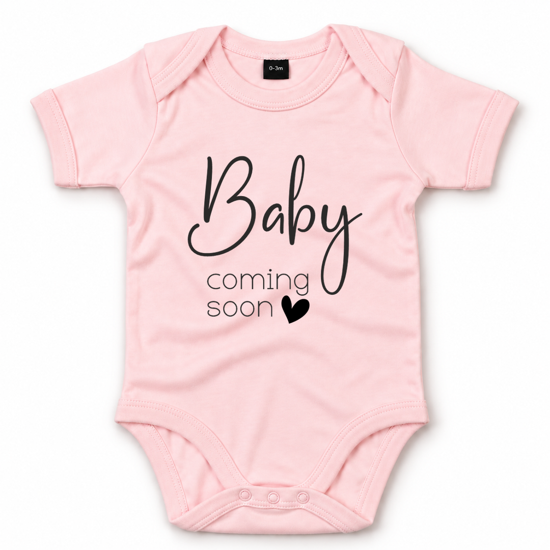 Roze zwangerschapsaankondiging romper met zwarte tekst Baby Coming Soon.