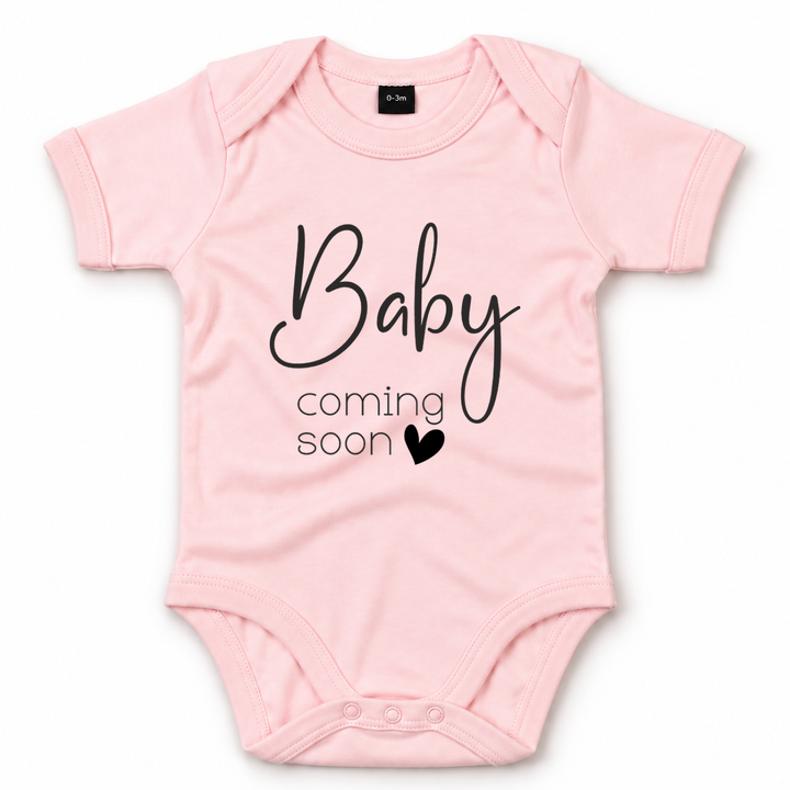 Roze zwangerschapsaankondiging romper met zwarte tekst Baby Coming Soon.